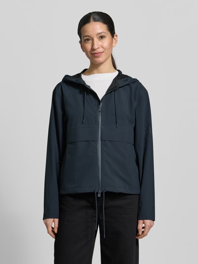Modelka nosi Regular Fit Kurtka z kapturem Model 'Suva' od Rains; zamek błyskawiczny dwustronny, tunel w pasie; ciemny kolor, gładka tekstura. Ganzkörperansicht von vorne. Długość tyłu w rozmiarze S: 59 cm, szerokość ramion w rozmiarze S: 49 cm, szerokość w rozmiarze S: 54 cm, długość rękawów w rozmiarze S: 60 cm.