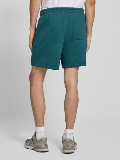 Męski model nosi Relaxed Fit Shorts mit Logo-Stitching von New Balance z mieszanki bawełny, kolor zielony, elastyczny pas z tunelem, szerokość w rozmiarze M: 42 cm, długość wewnętrzna nogawki: 18 cm, widok całej sylwetki z tyłu.