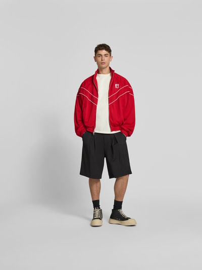Männermodel trägt Bomberjacke mit Raglanärmeln von RHUDE in Rot mit Stehkragen, Zweiwege-Reißverschluss, Reißverschlusstaschen, lockere Passform, schwarze Shorts, hellem Oberteil, schwarze Socken und beigefarbene Sneaker, glatte Struktur, Ganzkörperansicht von vorne. Rückenlänge 61 cm, Schulterbreite 49 cm, Ärmellänge 67 cm in Größe M.
