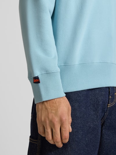 Männermodel trägt Sweatshirt mit Rundhalsausschnitt und Label-Stitching von Superdry in Hellblau, Baumwoll-Mix, Nahaufnahme Oberkörper. Regular-Fit, Rückenlänge bei Größe M 61 cm, Ärmellänge 64 cm.