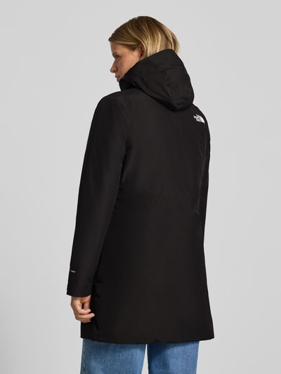 Modelka kobieca nosi czarną kurtkę z kapturem, zamek błyskawiczny dwustronny, Kieszenie zapinane na zamek błyskawiczny, Model 'HIKESTELLER' od The North Face, krój regular fit, Logo, Długość tyłu w rozmiarze S 86 cm, Szerokość ramion w rozmiarze S 41 cm, Szerokość w rozmiarze S 52 cm, Długość rękawów w rozmiarze S 65 cm, widok całej sylwetki z tyłu.