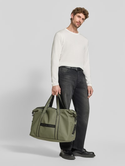 Männermodel trägt Weekender mit Logo-Detail Modell 'Däsh' von GASTON LUGA in Khaki, schwarze Details, Ganzkörperansicht von vorne. Hauptfach mit Reißverschluss, Schulterriemen 62 cm, Maße 64x40x25 cm.