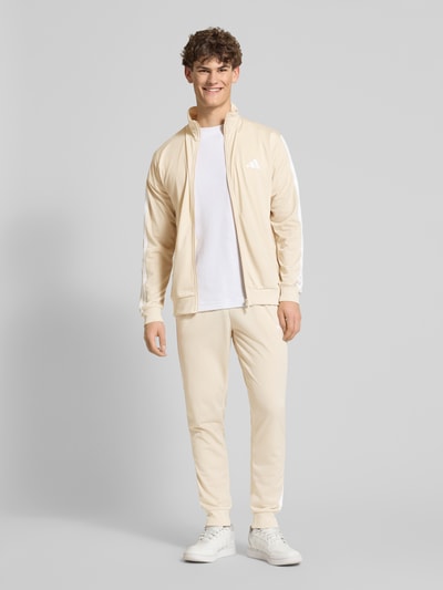 Männermodel trägt Regular Fit Sweat-Anzug mit Kontraststreifen von adidas Sportswear in Beige, Reißverschlussjacke, seitliche Taschen, Ganzkörperansicht von vorne.