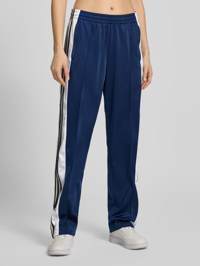Frauenmodel trägt Straight Fit Jogginghose mit elastischem Bund Modell 'Adibreak' von adidas Originals in Dunkelblau mit weißen Ziernähten und seitlichen Streifen, Taschen sichtbar, Ganzkörperansicht von vorne.