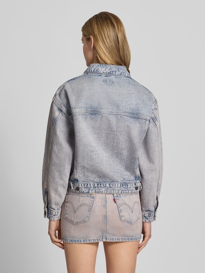 Frauenmodel trägt Oversized Jeansjacke mit Brusttaschen und Label-Patch von Guess Jeans in hellem Denim mit Label-Patch, Baumwoll-Mix mit Stretch-Anteil, Oversized-Fit, Kentkragen, Knopfleiste, seitlichen Eingrifftaschen, Rückansicht Ganzkörperansicht.