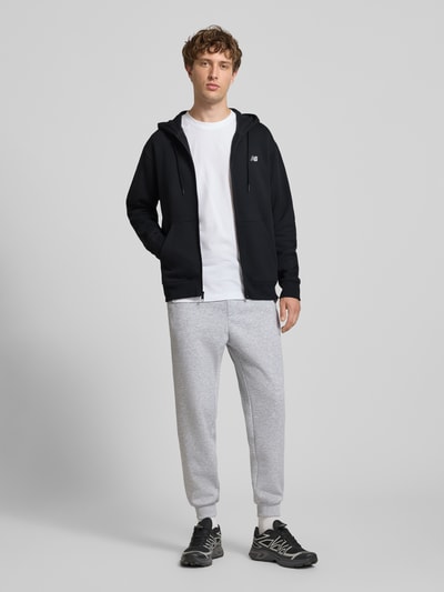 Männermodel trägt schwarze Sweatjacke mit Känguru-Tasche und Label-Stitching von New Balance, Baumwoll-Mix, Ganzkörperansicht von vorne
