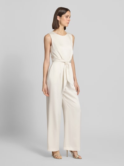 Modelka kobieca nosi Jumpsuit aus Satin mit Bindegürtel von Christian Berg Cocktail w kolorze kremowym, krój lekko taliowany, okrągły dekolt, pasek na wysokości talii, tylny zamek, proste nogawki, gładka satynowa tekstura, Szerokość w rozmiarze 36: 42 cm, długość całkowita 155 cm, długość wewnętrzna nogawki 81 cm, widok całej sylwetki z przodu.