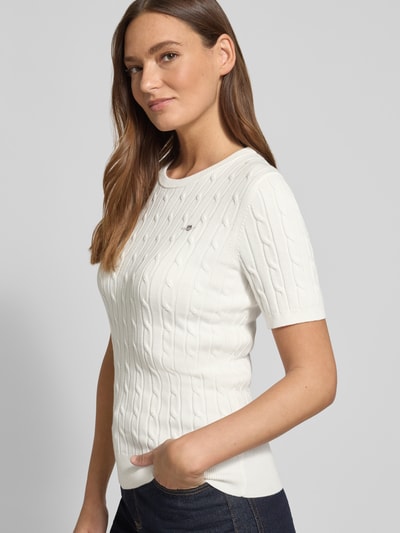 Modelka nosi Strickshirt mit Zopfmuster und Label-Stitching od Gant, mieszanka bawełny z dodatkiem streczu, krój slim fit, okrągły dekolt, detal ze ściegiem warkoczowym, wyhaftowane logo, kolor biały, długość tyłu w rozmiarze S 57 cm, długość rękawa w rozmiarze S 24 cm, widok na cały korpus z przodu.