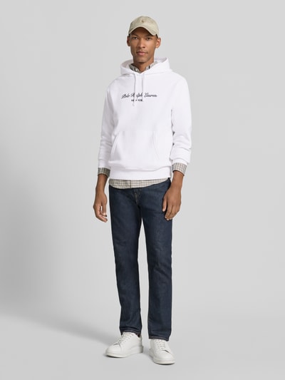 Mannelijk model draagt Relaxed Fit Hoodie uit katoenmix van Polo Ralph Lauren in wit met logo, donkere jeans, lichte sneakers, Ganzkörperansicht van voren.