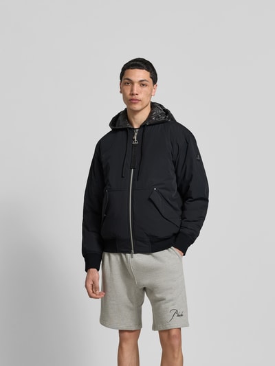 Männermodel trägt schwarze Bomberjacke mit Daunen-Feder-Füllung von Moose Knuckles, lockere Passform, Kapuze, Känguru-Tasche, Zweiwege-Reißverschluss, Ganzkörperansicht von vorne. Rückenlänge 63 cm, Schulterbreite 48 cm, Ärmellänge 65 cm bei Größe M.