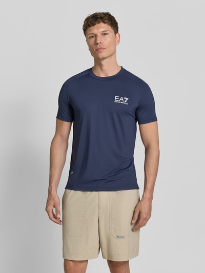 Männermodel trägt T-Shirt mit Label-Print von EA7 Emporio Armani in Dunkelblau, Slim Fit mit Rundhalsausschnitt, Ganzkörperansicht von vorne.