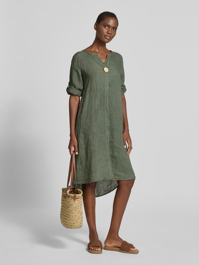 Kobiece model nosi knielanges Leinenkleid mit Zierknopf Modell 'Ad44ora' von ZABAIONE z lnu, linia a, długość do kolan, dekolt w serek, ozdobny guzik, rękaw o dł. 3/4, długość tyłu w rozmiarze S: 107 cm, szerokość w rozmiarze S: 47 cm, długość rękawów w rozmiarze S: 30 cm; matowa, oliwkowozielona tkanina, struktura lekko marszczona. Ganzkörperansicht z przodu.