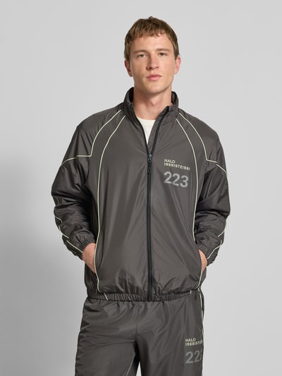 Männermodel trägt Trainingsjacke mit Reißverschlusstaschen von HALO in Grau mit Label-Print, Regular Fit, Stehkragen, Ganzkörperansicht von vorne.