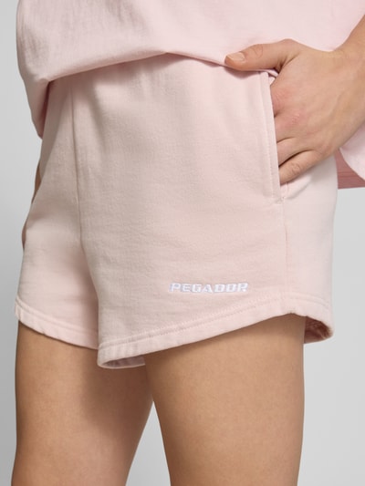 Kobieta nosi szorty z wysokim stanem High Waist Shorts mit Logo-Stitching model 'WILUNA' od Pegador z mieszanki bawełny, wpuszczane kieszenie, elastyczny pas, wyhaftowane logo, szerokość w rozmiarze S: 40 cm, długość wewnętrzna nogawki w rozmiarze S: 7 cm, detaliczna faktura, kolor jasnoróżowy, zbliżenie na biodra i wpuszczaną kieszeń, detaliczny kadr.