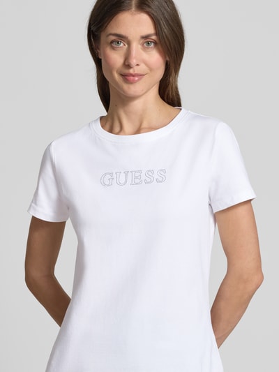 Frauenmodel trägt T-Shirt mit Logo Modell 'BRIANA' von Guess Activewear aus weißem Baumwoll-Elasthan-Mix, Logo mit Ziersteinbesatz, Nahaufnahme Oberkörper.