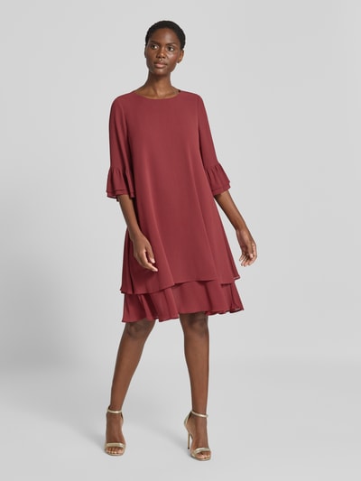 Frauenmodel trägt Relaxed Fit Kleid in Knielänge von Christian Berg Cocktail in Rot mit Rüschenbesatz, Halbarm, fließende Struktur, Ganzkörperansicht von vorne.