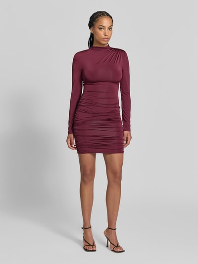 Frauenmodel trägt Slim Fit Minikleid aus Viskose-Mix von ROTATE in Burgunderrot mit Raffungen, Stehkragen und langen Ärmeln, Ganzkörperansicht von vorne. Gesamtlänge 85 cm, Brustweite 32 cm, Ärmellänge 64 cm.