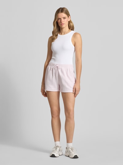 Frauenmodel trägt Straight Fit Shorts mit Tunnelzug Modell 'French' von New Balance aus Baumwoll-Mix in zartem Rosa, Ganzkörperansicht von vorne. Damen Shorts, elastischer Bund, Bundweite 40 cm, Innenbeinlänge 8 cm, Größe S.