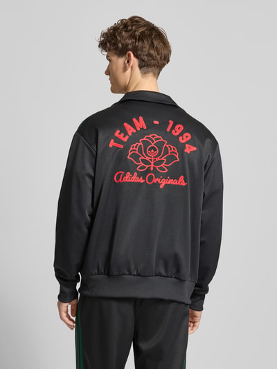 Männermodel trägt Sweatjacke mit Logo-Stitching und Stehkragen van adidas Originals, materiaal-mix met katoen, rechte fit, elastische boorden, rits en borduring, zwarte stof met rode accenten, rugaanzicht Ganzkörperansicht Rückansicht.