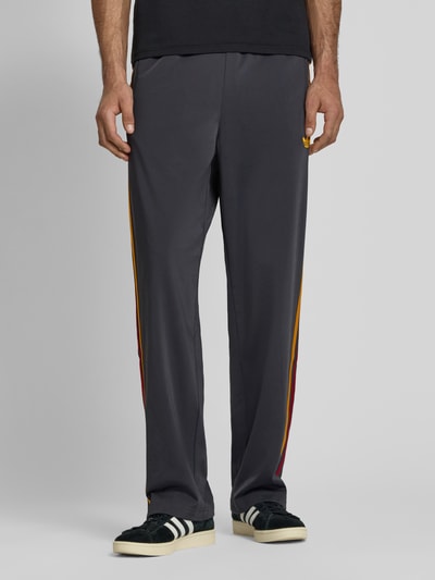 Mężczyzna nosi Relaxed Fit Sweatpants mit Label-Streifen Modell 'FIREBIRD' od adidas Originals, efekt: jednolity kolor, detal z logo, paski z logo, elastyczny ściągacz, tunel, zakończenia nogawek z zamkiem błyskawicznym, boczne kieszenie zapinane na zamek błyskawiczny, wykończenie materiału: dzianina dresowa, Ganzkörperansicht z przodu.