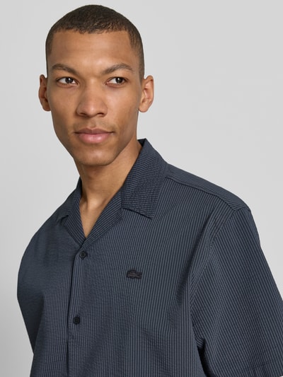 Mężczyznę model nosi Relaxed Fit Freizeithemd aus reiner Baumwolle von Lacoste, tkanina stylizowana na denim, marszczenie, naszywka z logo, krótki rękaw, kołnierzyk koszulowy, listwa guzikowa na całej długości, bawełna, zbliżenie na górną część ciała.
