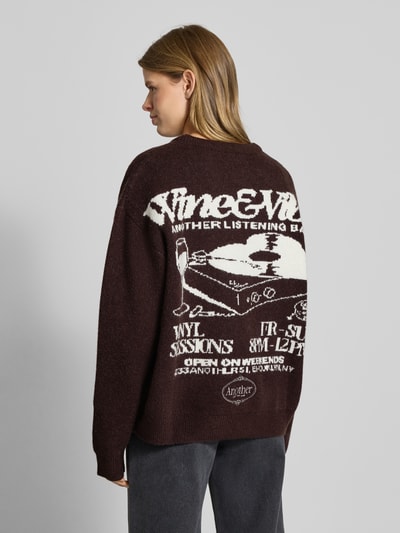 Frauenmodel trägt Oversized Strickpullover mit Rückenmotiv Modell 'Wine & Vibe' von ANOTHER COTTON LAB, Rundhalsausschnitt, Label-Stitching, Rückenlänge bei Größe S 62 cm, Ärmellänge 68 cm, dunkle Farbe, grobe Strickstruktur, Ganzkörperansicht Rückansicht.