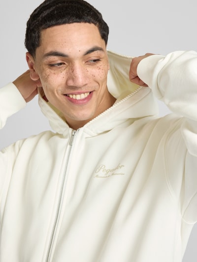 Männermodel trägt Oversized Sweatjacke mit Känguru-Tasche Modell 'Morcote' von Pegador aus Baumwoll-Mix in Creme, Nahaufnahme Oberkörper. Rückenlänge 72 cm, Ärmellänge 65 cm, Kapuze und Reißverschluss.