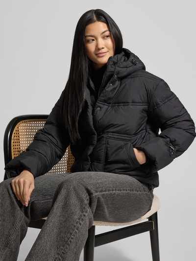 Frauenmodel trägt Cropped Fit Steppjacke mit aufgesetzten Taschen Modell 'Daisy' von Guess, Schwarz, Allover-Logo-Print, Gefütterte Kapuze, Nahaufnahme Oberkörper.