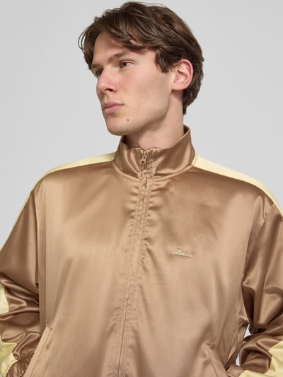 Männermodel trägt Schimmernde Trainingsjacke mit Label-Stitching von BLKVIS in Braun mit gelben Details, Stehkragen, Reißverschluss, Nahaufnahme Oberkörper. Rückerlänge bei Größe M: 61 cm, Ärmellänge: 63 cm, Regular-Fit, schimmernde Textur.
