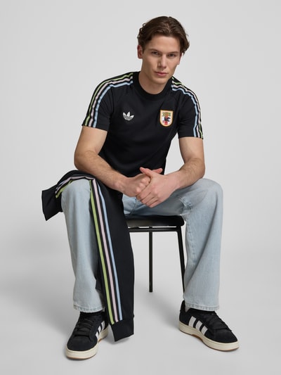 Männermodel trägt Regular Fit T-Shirt aus Baumwoll-Mix mit JFA-Applikation Modell 'NOIR' von adidas Originals, unifarben, Kurzarm, Rundhals, Logo-Stitching und Kontraststreifen, dazu helle Jeans und Sneaker, Ganzkörperansicht von vorne.