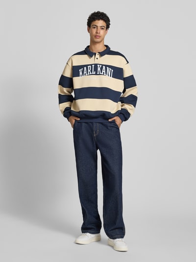 Männermodel trägt Oversized Sweatshirt mit Polokragen Modell 'Heritage' von KARL KANI in Beige und Dunkelblau, Baumwoll-Mix, Logo-Print, Elastische Abschlüsse, Rückenlänge bei Größe M 65 cm, Ganzkörperansicht von vorne.