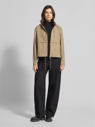 Frauenmodel trägt Regular Fit Jacke mit Kapuze Modell 'Suva' von Rains in Beige mit Tunnelzug, Zweiwege-Reißverschluss, Ganzkörperansicht von vorne.