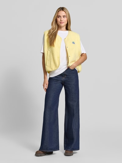 Frauenmodel trägt Relaxed Fit Weste aus Teddyfell Modell 'Hoktown' von American Vintage in Gelb mit Logo-Patch und Knopfleiste, Ganzkörperansicht von vorne. Rückenlänge 56 cm, Schulterbreite 43 cm, Brustweite 63 cm.