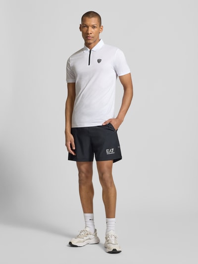 Męski model nosi Slim Fit Poloshirt aus Viskose-Mix od EA7 Emporio Armani, biały, kołnierzyk polo, detal z logo, elastyczna mieszanka wiskozy, krój slim fit, elastyczne zakończenia rękawów, długość tyłu w rozmiarze M: 60 cm, widok całej sylwetki z przodu.