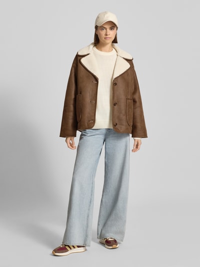 Modelka nosi kurtkę Jacke in Leder-Optik mit Futter aus Teddyfell od Review w kolorze brązowym, krój regular fit, kołnierz z połami, listwa guzikowa, nakładane kieszenie, podszewka z futerka, długość tyłu w rozmiarze S: 67 cm, szerokość ramion w rozmiarze S: 52 cm, szerokość klatki piersiowej w rozmiarze S: 60 cm, długość rękawów w rozmiarze S: 55 cm; całkowity widok od przodu.