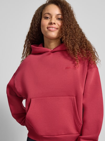 Modelka kobieca nosi Regular Fit Hoodie mit Kängurutasche od Review, bawełniana mieszanka, krój regular fit, kieszeń kangurka, kaptur, detal z logo, elastyczne wykończenia, zbliżenie na górną część ciała.
