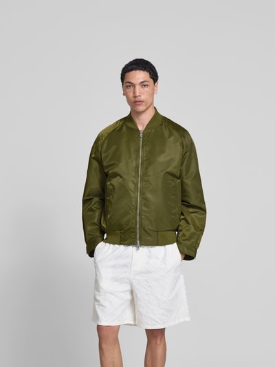 Männermodel trägt Bomberjacke mit Stehkragen von Filippa K in Olivgrün, Relaxed-Fit, Eingrifftaschen, Zweiwege-Reißverschluss, elastischer Bund, Ganzkörperansicht von vorne. Rückenlänge 57 cm, Schulterbreite 46 cm, Ärmellänge 67 cm bei Größe 50.