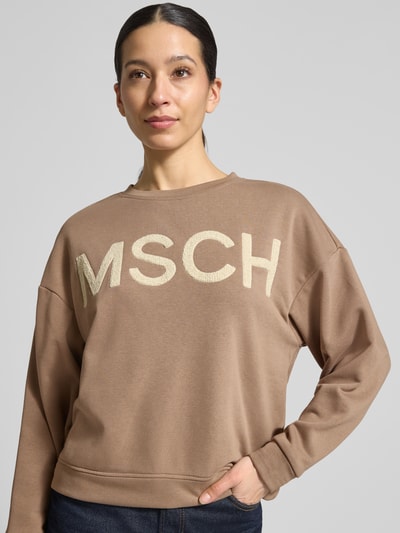 Modelka kobieca ma na sobie Sweatshirt mit überschnittenen Schultern od MSCH Copenhagen, bawełniany miks, prosty krój, model 'Mela Briena', beżowy kolor, tekstura gładka, widoczne duże logo, widok na górną część ciała z przodu.