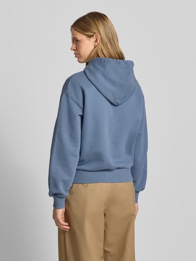Frauenmodel trägt Oversized Hoodie aus Baumwoll-Mix Modell 'CASEY' von Carhartt Work In Progress, wyhaftowane logo, mieszanka bawełny i elastanu, krój oversized, kaptur z tunel, prążkowane zakończenia rękawów, kieszeń kangurka, jasnoniebieska bluza, beżowe spodnie, matowa tekstura, całkowity widok z tyłu