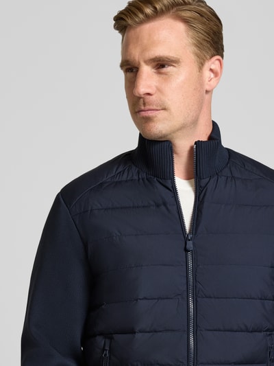 Männermodel trägt Steppjacke mit geripptem Stehkragen Modell 'SEDUM' von Save The Duck in Dunkelblau, Nahaufnahme Oberkörper, gerader Schnitt, Strickärmel, Reißverschlusstaschen, Label-Patch.