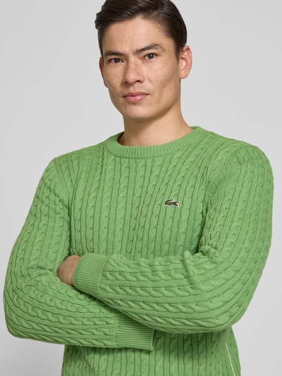 Männermodel trägt Regular Fit Strickpullover aus Baumwoll-Mix von Lacoste in Grün, Optik: Unifarben, Zopfmuster, Logo-Patch, Nahaufnahme Oberkörper.