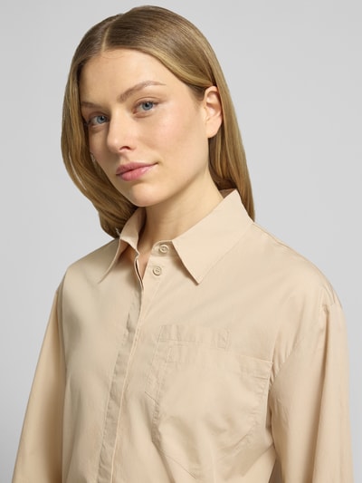 Kobiece model prezentuje Boxy Fit Bluse mit Brusttaschen von Marc Cain, beżowa bawełna, kołnierzyk koszulowy, listwa guzikowa, brustasza, mieszanka gładkich tekstur, długość tyłu 51 cm, długość rękawów 57 cm, zbliżenie na górną część ciała.