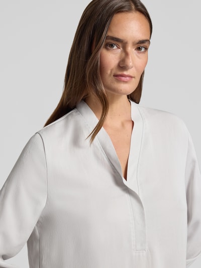 Kobieta modelka nosi Regular Fit Bluse mit V-Ausschnitt von Marc Cain, kolor jasnoszary, detal z logo, długość tyłu 65 cm, długość rękawów 59 cm, krój regular fit, dekolt w serek, bluzka damska z długim rękawem, detal z logo, Nahaufnahme górnej części ciała.