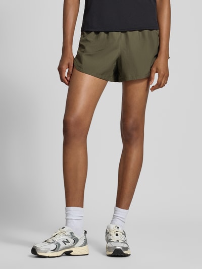Frauenmodel trägt Shorts mit elastischem Bund Modell 'NAGINO' von ASICS TIGER in Olive, strukturierte Oberfläche, seitliche Taschen, Nahaufnahme Oberkörper bis Oberschenkel, Kontext: gerade Passform, reflektierende Details, Reißverschlusstasche hinten, Bundweite Größe S 34 cm, Innenbeinlänge 14 cm.