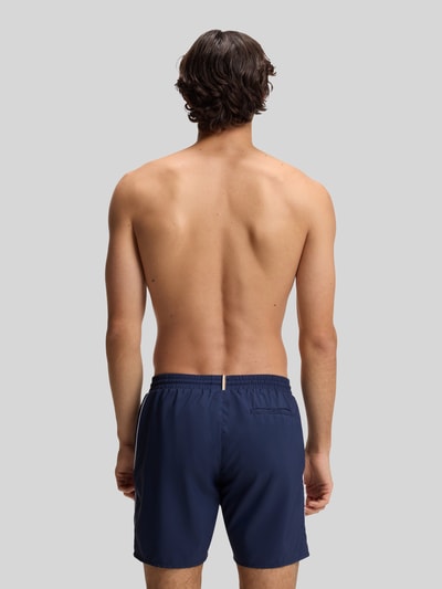 Männermodel trägt blaue Badeshorts mit Kontrast-Logo Modell 'STARFISH' von BOSS, elastischer Bund, unifarbene Optik, Ganzkörperansicht Rückansicht.