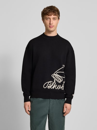 Männermodel trägt Strickpullover mit Label-Detail von BLKVIS in Schwarz, lockerer Schnitt, reine Baumwolle, Rundhalsausschnitt, gerippte Abschlüsse, Rückenlänge 68 cm, Ärmellänge 60 cm, Ganzkörperansicht von vorne.