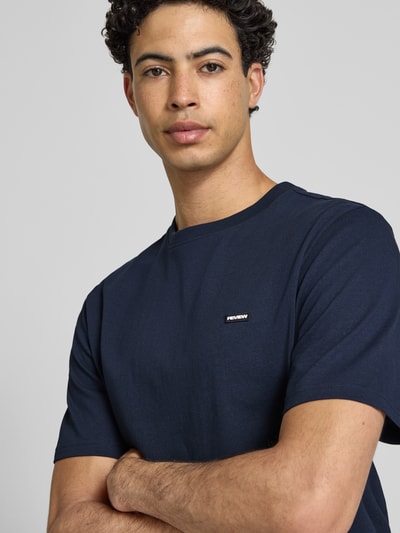 Männermodel trägt T-Shirt mit Label-Patch und Rundhalsausschnitt von REVIEW, Reine Baumwolle, Boxy-Fit, Nahaufnahme Oberkörper.