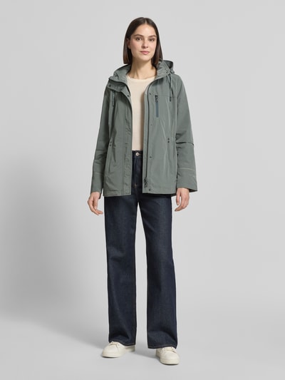 Frauenmodel trägt Funktionsjacke mit Brusttasche von Gil Bret in Grün, mit Kapuze, Stehkragen, Tunnelzug, Tailliert Passform, Ganzkörperansicht von vorne. Rückenlänge bei Größe 36: 74 cm, Schulterbreite: 41 cm, Brustweite: 47 cm, Ärmellänge: 65 cm.