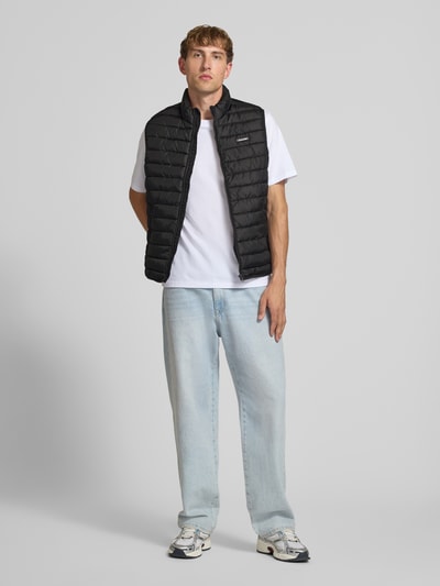 Männermodel trägt Steppweste mit Reißverschlusstaschen Modell 'ELVIS' von JACK & JONES in Schwarz, kombiniert mit weißem Shirt und Jeans, Ganzkörperansicht von vorne.