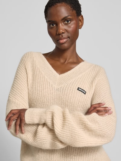 Frauenmodel trägt Oversized Pullover mit Woll-Anteil von ROTATE in Beige, Strick-Optik, V-Ausschnitt, Logo-Patch, Material-Mix, Nahaufnahme Oberkörper.